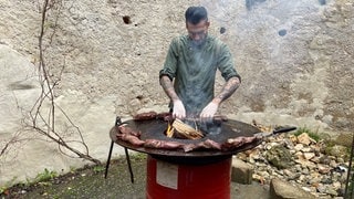 Ein Koch steht an einem Metallfass. Darin brennt ein Feuer. Auf einer Eisenplatte am oberen Fassrand grillt er Fleisch.