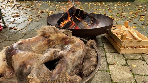 Im Vordergrund sind Tierhäute in einer Schale zu sehen, in Hintergrund brennt ein Feuer in einer Schale. 