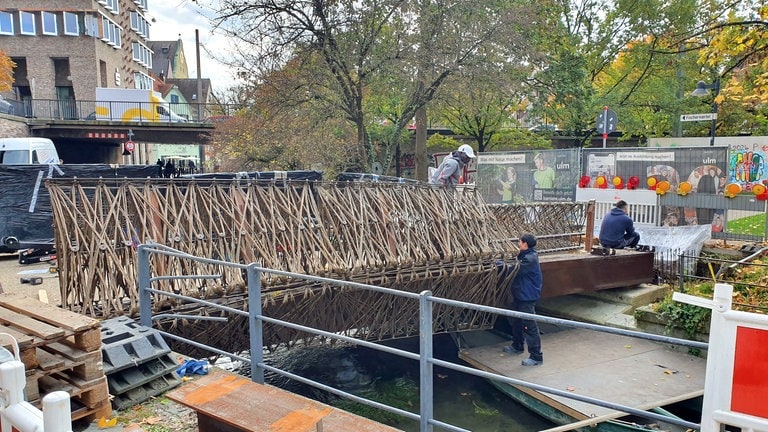 So langsam nimmt die viel diskutierte Fußgängerbrücke über die Blau in der Ulmer Innenstadt Gestalt an: Am Dienstag wurde mit der Montage der Geländer der Öko-Brücke begonnen, auch sie bestehen aus Flachsfasern und Bio-Harz. Für die Stadt Ulm ist die "Smart Circular Bridge" ein ökologisches Vorzeigeprojekt, für den Bund der Steuerzahler wegen der stark gestiegenen Kosten ein Fall von Steuerverschwendung (22.10.).