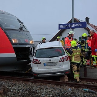 Zug und Auto nach einem Zusammenstoß, Rettungskräfte darum herum: Vom Zug erfasst, halb auf dem Bahnsteig: Ein 18-jähriger Autofahrer ist am Donnerstagmorgen bei Aletshausen im Kreis Günzburg mit einer Regionalbahn zusammengestoßen.