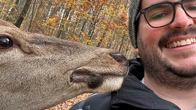 Hirschkuh Gretl vom Wildpark Heidenheim begutachtet ganz nah die Wange des SWR Reporters Dennis Bechtold, der in die Kamera lächelt. 