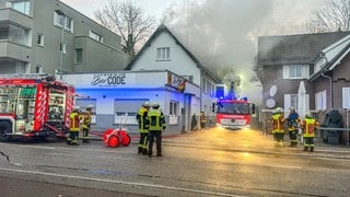 Rauch dringt aus dem Dachstuhl eines Hauses - auf der Straße davor stehen Feuerwehrleute mit ihrem Einsatzfahrzeug. 