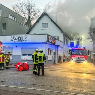 Rauch dringt aus dem Dachstuhl eines Hauses - auf der Straße davor stehen Feuerwehrleute mit ihrem Einsatzfahrzeug. 