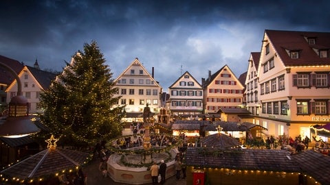 Umgeben von beleuchteten Giebeln ist der Biberacher Christkindles-Markt auf dem mittelalterlichen Marktplatz zu sehen. Die kleinen Holzbuden, zwischen denen Besucherinnen und Besuchern bummeln, sind festlich geschmückt und in der Mitte steht ein großer Tannenbaum mit Lichterketten.