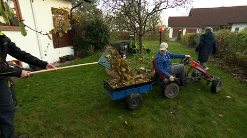 Ein Kettcar samt Anhänger steht im Garten eines Wohnhauses. Mehrere Kinder rechen Laub zusammen und laden es in den Anhänger.