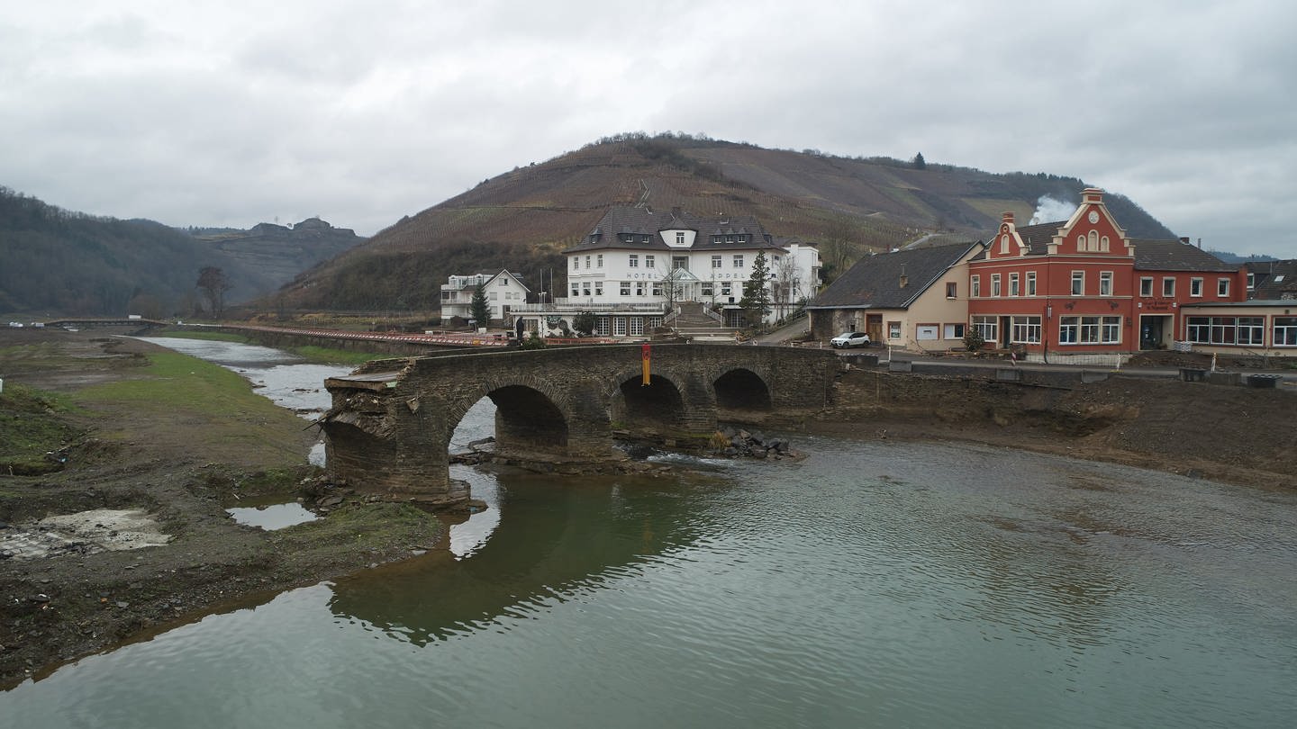 Eine zerströrte Brücke steht ohne Verbindung zu den Flussufern in der Ahr - im Hintergrund sind noch immer unterschiedlich stark von Flutschäden betroffene Gebäude zu erkennen.