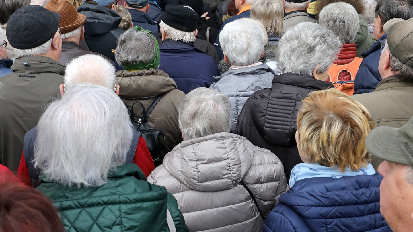 Eine Große Gruppe alter Menschen in Winterjacken steht an, um Einlass in eine Veranstaltungshalle zu bekommen.