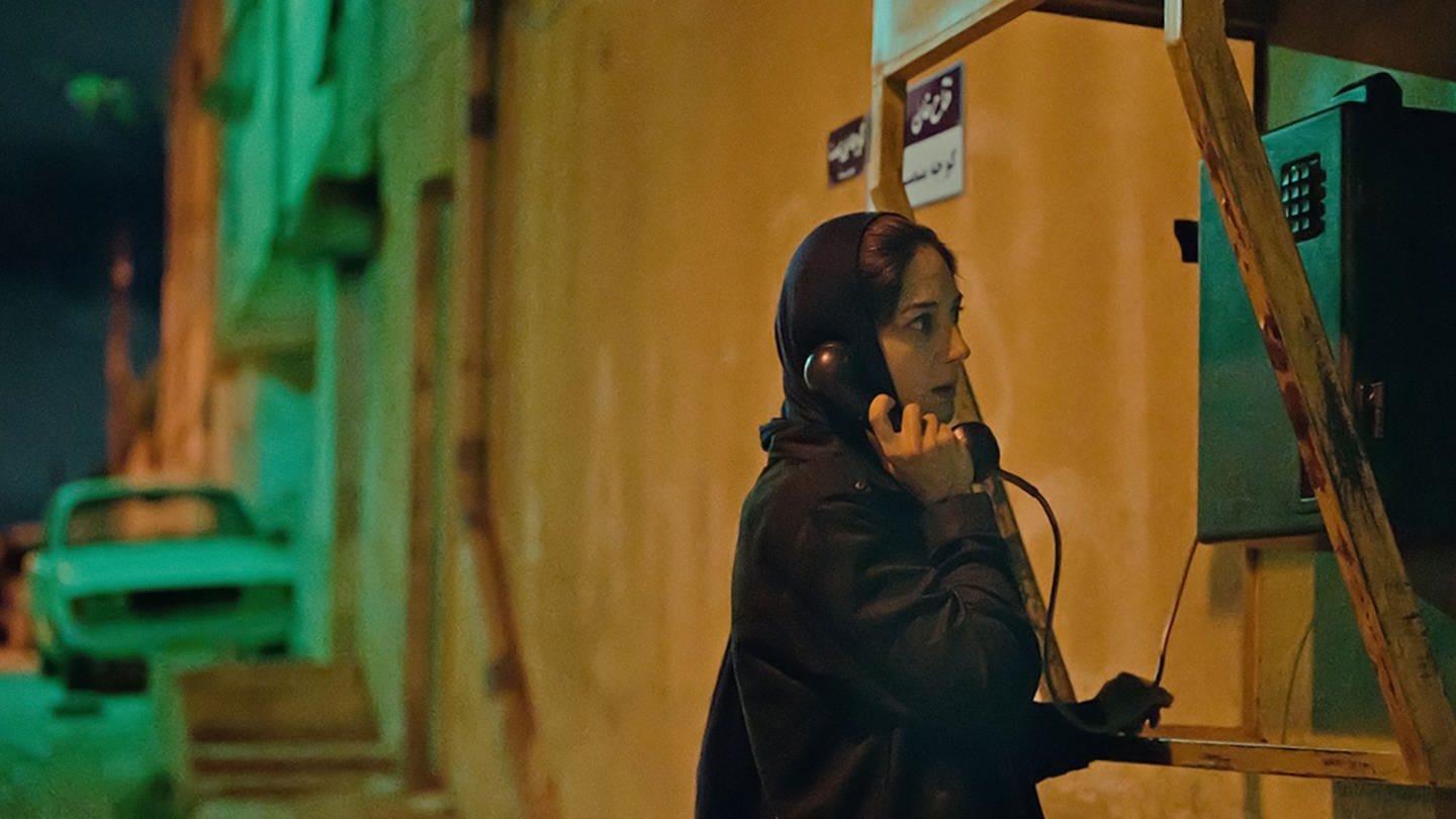 In der Filmszene ist eine junge Frau zu sehen, die bei Nacht in einer dunklen Gasse ein öffentliches Telefon benutzt.