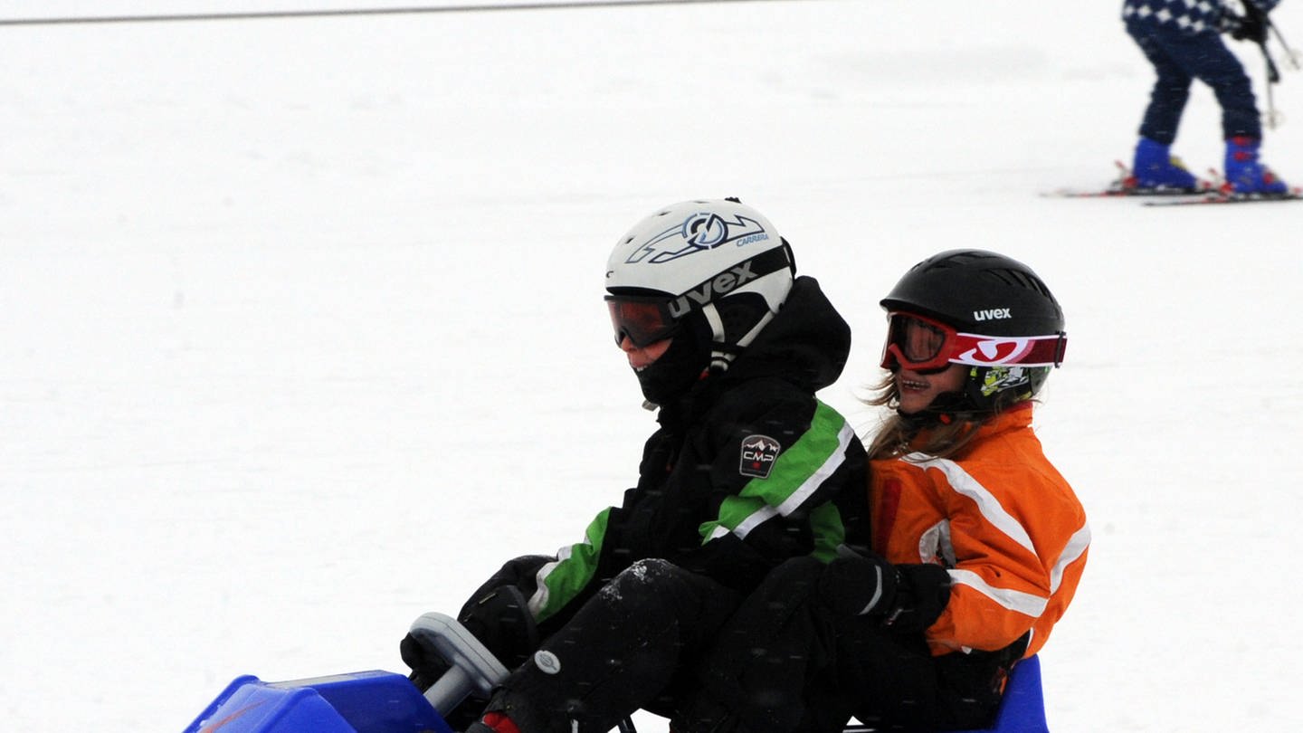 Zwei Kinder rutschen auf einem blauen Schlitten eine Skipiste herunter