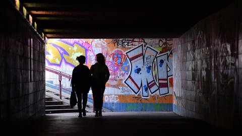 Menschen laufen durch eine dunkle Unterführung, im Hintergrund bunte Graffiti.