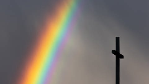 Ein kräftiger Regenbogen ist am dunklen Himmel neben dem Kreuz einer Kirche zu sehen.