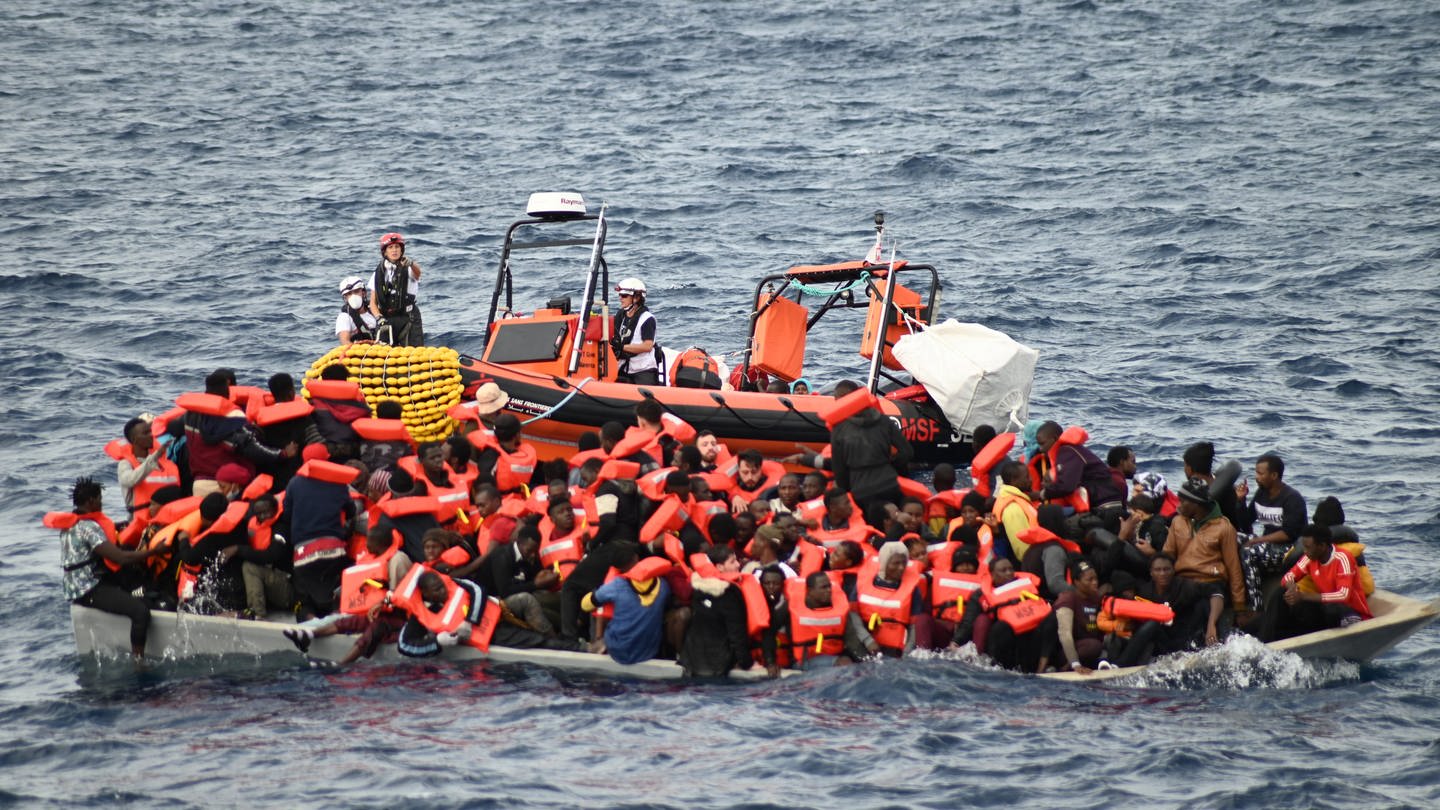 Ein Rettungsboot schwimmt hinter einem mit Migranten besetzten Boot.