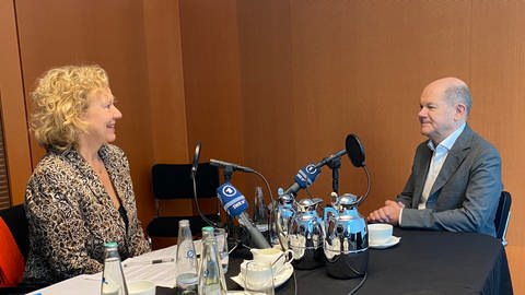 SWR Korrespondentin Evi Seibert und Kanzler Olaf Scholz sitzen sich beim Interview an einem Tisch mit Mikrofonen und Getränken gegenüber