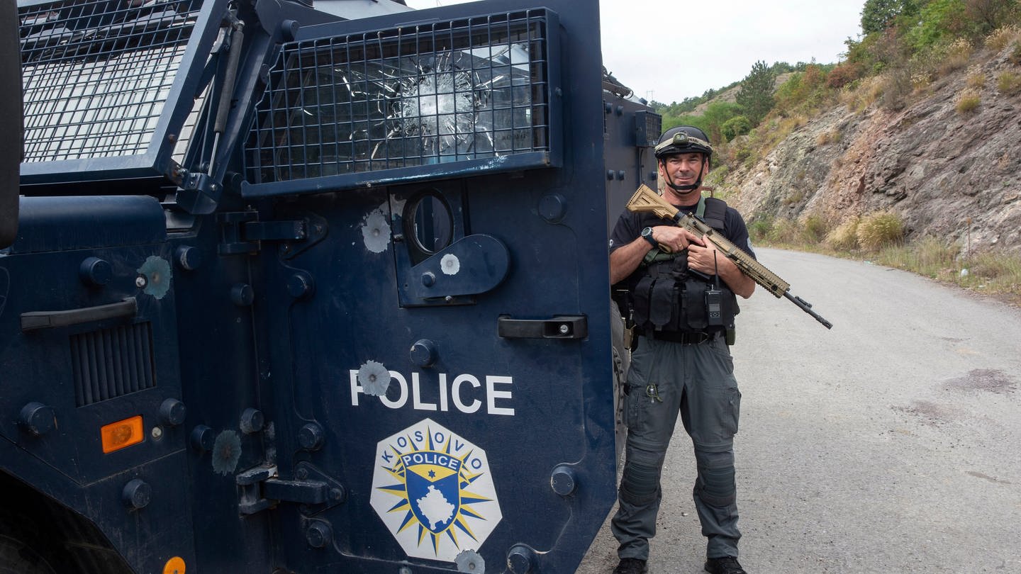 Ein Polizist steht im Kosovo neben einem Panzerwagen, er hält ein Sturmgewehr schräg vor der Brust und grinst