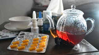 Medikamente und eine Kanne und Tasse Tee zur Bekämpfung einer Erkältung sind auf einem Nachttisch aufgestellt.