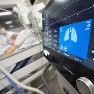 Eine Pflegerin versorgt auf einer Intensivstation eine Patientin, im Vordergrund ist der Monitor eines Beatmungsgeräts zu sehen