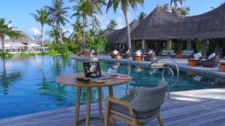 Ein Arbeitsplatz mit Laptop steht auf einem Café-Tisch mit Sessel neben einem Pool, umringt mit Palmen und Hütten einer Hotel-Anlage auf den Malediven.