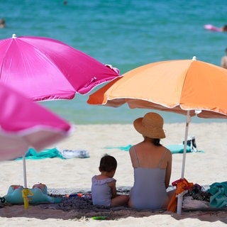 Eine Frau sitzt mit einem Kind unter dem Schatten von zwei Sonnenschirmen am Strand und blickt aufs Mittelmeer.