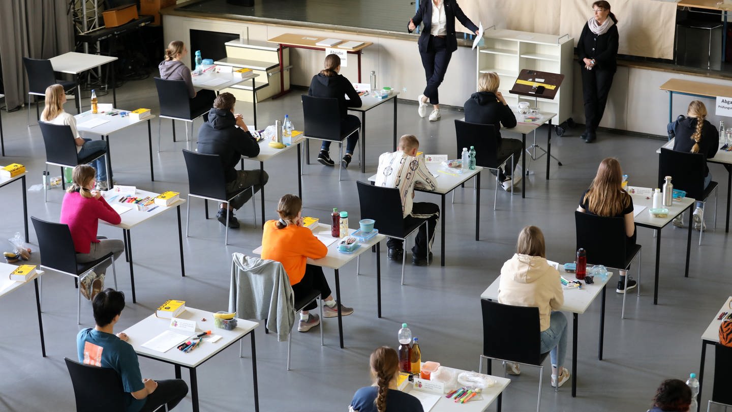 In der Aula in einem Gymnasium beginnt die schriftliche Abiturprüfung in Deutsch - Schülerinnen und Schüler sitzen an Einzeltischen und werden beaufsichtigt.