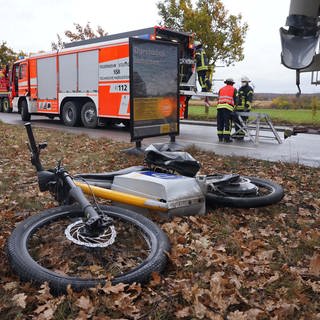 Ein E-Bike liegt nach einem Unfall hinter einer Bushaltestelle auf einer Wiese. Im Hintergrund sieht man ein Feuerwehrauto und Feuerwehrmänner.