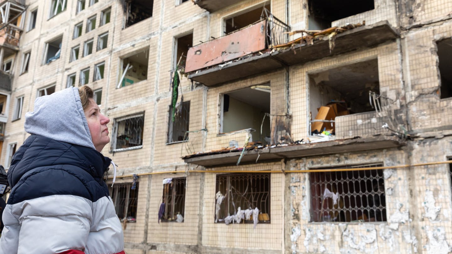 Eine alte Frau steht vor einer Ruine eines Mehrfamilienhauses in der Ukraine