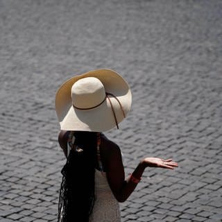 Eine Frau schützt sich mit einem Hut vor der sengenden Sonne, während die Temperaturen bis zu 39 Grad Celsius erreichen. Sie steht auf einem asphaltierten Platz.