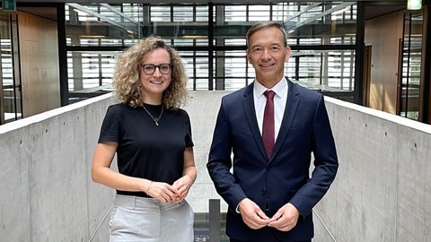 Korrespondentin Lissy Kaufmann und Pascal Kober stehen in der Halle im ARD-Hauptstadtstudio nebeneinander und lächeln in die Kamera