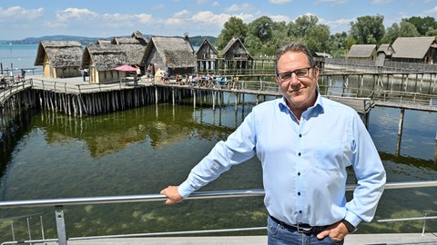 Professor Gunter Schöbel, Direktor des Pfahlbauten-Museums Unteruhldingen, steht auf der Terasse seines Büros, während im Hintergrund die Pfahlbauten im Bodensee zu sehen sind. 