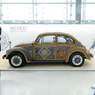 Ein VW-Käfer mit Bemalung in einem mexikanischen Ornament-Stil steht vor einer Wand mit der Aufschrift "México" in einer Ausstellungshalle.