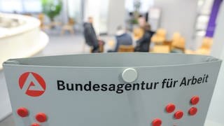 Drei Personen sitzen im Wartebereich der Bundesagentur für Arbeit hinter einem Aufsteller mit dem Logo der Agentur