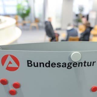 Drei Personen sitzen im Wartebereich der Bundesagentur für Arbeit hinter einem Aufsteller mit dem Logo der Agentur