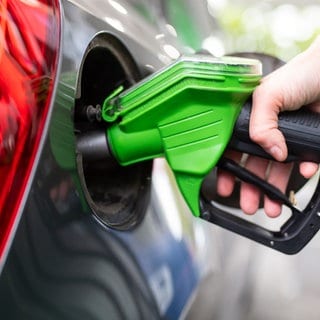 Eine Frau hält an einer Tankstelle an einer Zapfsäule eine Zapfpistole in der Hand und betankt ein Auto.