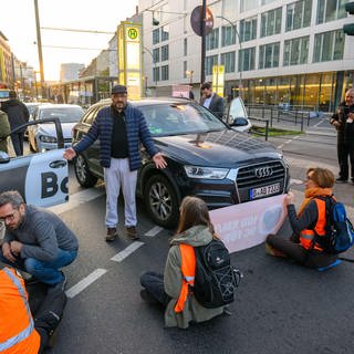 Aktivistin sitzen auf einer Straße und blockieren sie für Autofahrer. Die Autofahrer diskutieren mit den Aktivisten der "letzten Generation". 