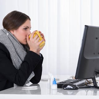 Eine Frau sitzt mit einem Schal und einer Tasse Tee vor einem Computer-Bildschirm.
