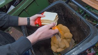 Eine Person holt Paprika, Kartoffeln und Toast aus einer Mülltonne. 