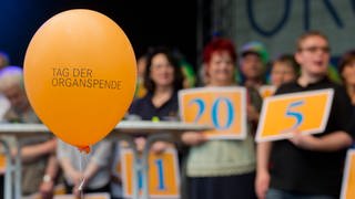 Ein Luftballon mit der Aufschrift "Tag der Organspende". Im Hintergrund Menschen, die Schilde hochalten, auf denen die Anzahl der Jahre steht, die sie nun mit Spenderorgan leben.