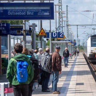 Ein ICE und ein Regionalzug  fahren an einem Bahnhof. Ziel ist laut Anzeigetafel das Ostseebad Binz. Dort soll es ab 2024 auch Direktverbindungen mit dem ICE aus Rheinland-Pfalz geben.