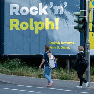 Frauen gehen an einem Plakat mit der Aufschrift "Rock 'n' Rolph!" vorbei, das Teil einer Mobilitäts-Kampagne der Landesregierung war.