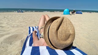 Sommer, Urlaub, Ferien: Eine Urlauberin mit Sonnenhut liegt am Sandstrand auf einem Handtuch und schaut auf das Meer.