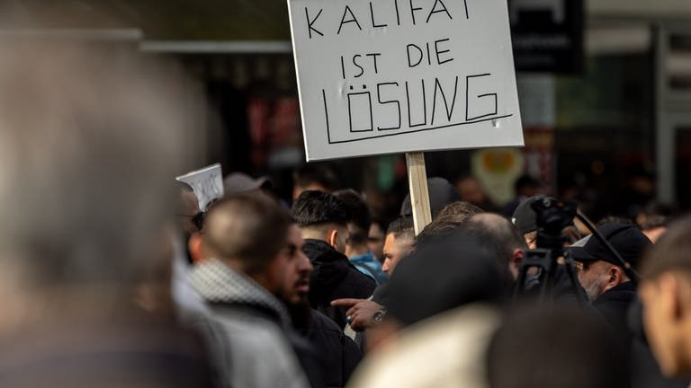 Teilnehmer einer Islamisten-Demo halten ein Plakat mit der Aufschrift „Kalifat ist die Lösung“ in die Höhe.