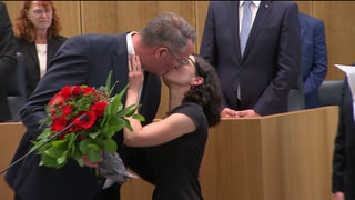 Der neue Ministerpräsident Alexander Schweitzer (SPD) wird nach seiner Wahl von seiner Frau mit einem Kuss beglückwünscht. Im Arm hält er einen roten Blumenstrauß. 
