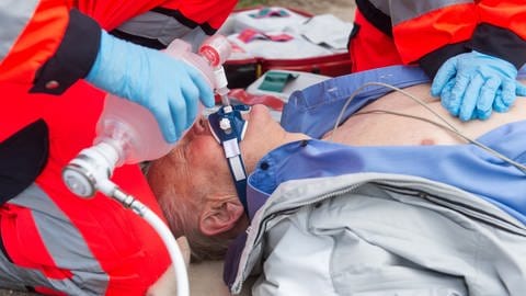 Ein Notarzt und ein Sanitäter versorgen einen Mann, der auf dem Boden liegt, mit Sauerstoff und führen eine Herzdruckmassage durch (gestellte Szene). 