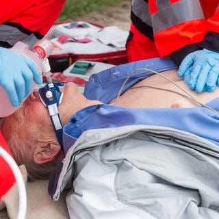 Ein Notarzt und ein Sanitäter versorgen einen Mann, der auf dem Boden liegt, mit Sauerstoff und führen eine Herzdruckmassage durch (gestellte Szene). 