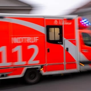 Ein rot-weißer Krankenwagen der Feuerwehr mit der Notrufnummer 112 fährt mit eingeschaltetem Blaulicht schnell durch die Stadt. 