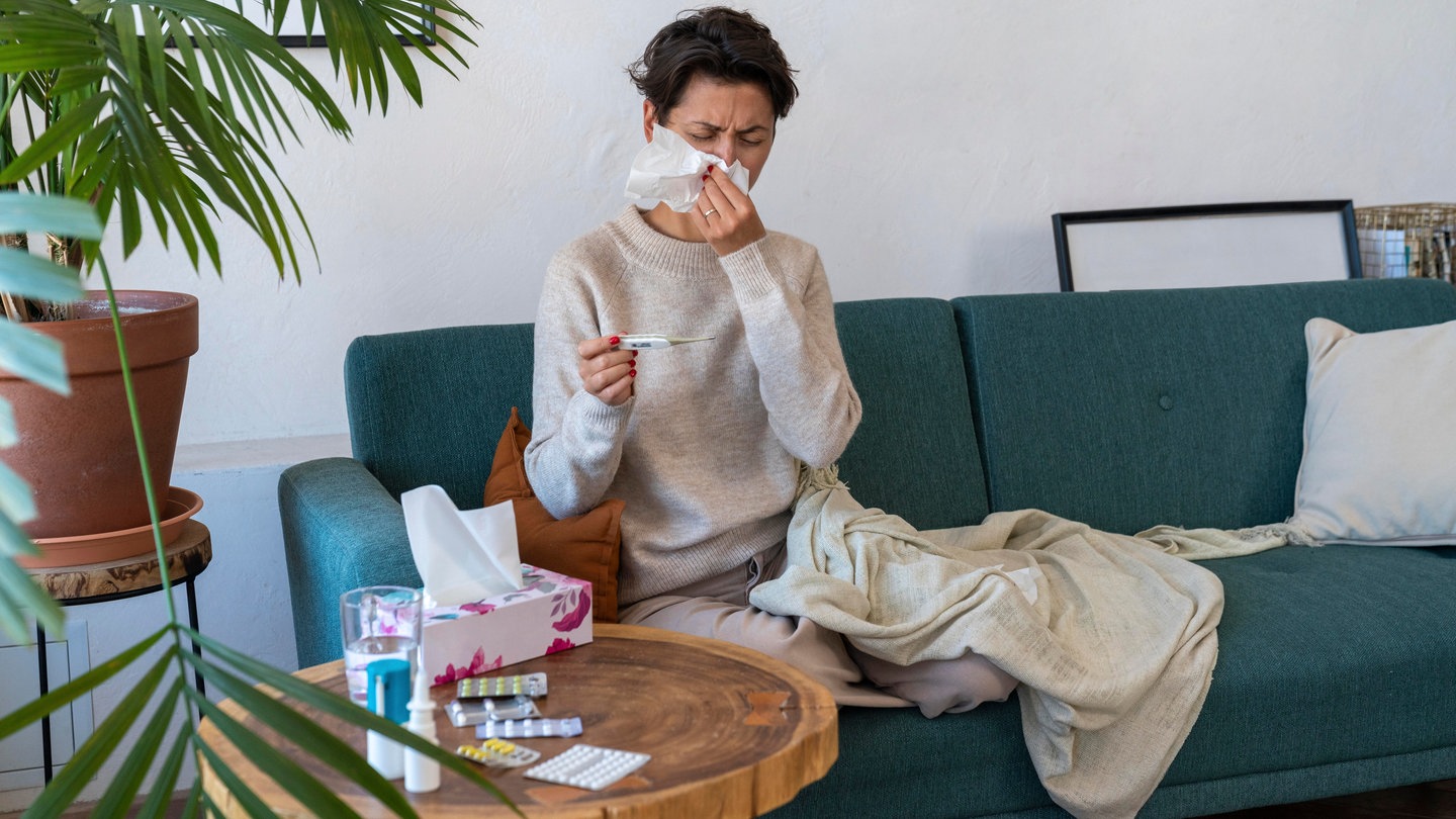 In RLP rollt die Erkältungswelle: Eine Frau sitzt abgeschlagen auf einem Sofa, schneuzt sich die Nase und schaut auf ein Fieberthermometer. Auf dem Tisch stehen Nasenspray, Nasendusche und Medikamente.