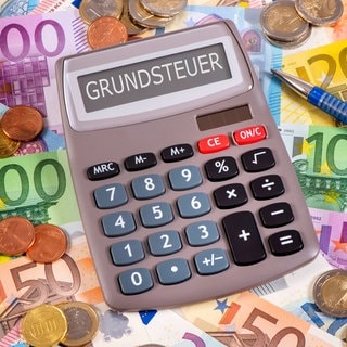 Ein Taschenrechner, auf dem "Grundsteuer" steht, liegt auf mehreren Geldscheinen. Die großen Städte in Rheinland-Pfalz planen nämlich wegen der Grundsteuer-Reform ihre Hebesätze zu erhöhen.