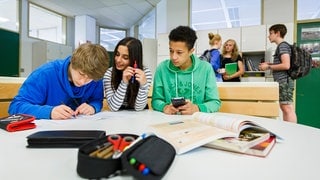 Zwei Schüler und eine Schülerin sitzen in einem Klassenraum an einem runden Tisch. Einer schreibt etwas, die anderen beiden gucken auf ihn.   