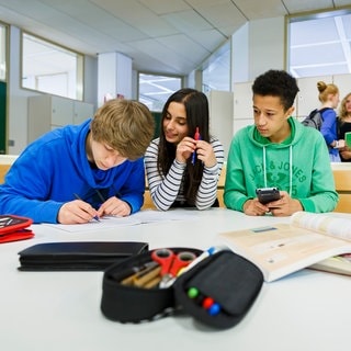 Zwei Schüler und eine Schülerin sitzen in einem Klassenraum an einem runden Tisch. Einer schreibt etwas, die anderen beiden gucken auf ihn.   