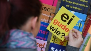 Eine Frau hält eine Postkarte mit dem klassischen hessischen Satz "Ei gude wie?" in der Hand. Jede Region hat ihre eigenen sprachlichen Einfärbungen.