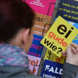 Eine Frau hält eine Postkarte mit dem klassischen hessischen Satz "Ei gude wie?" in der Hand. Jede Region hat ihre eigenen sprachlichen Einfärbungen.